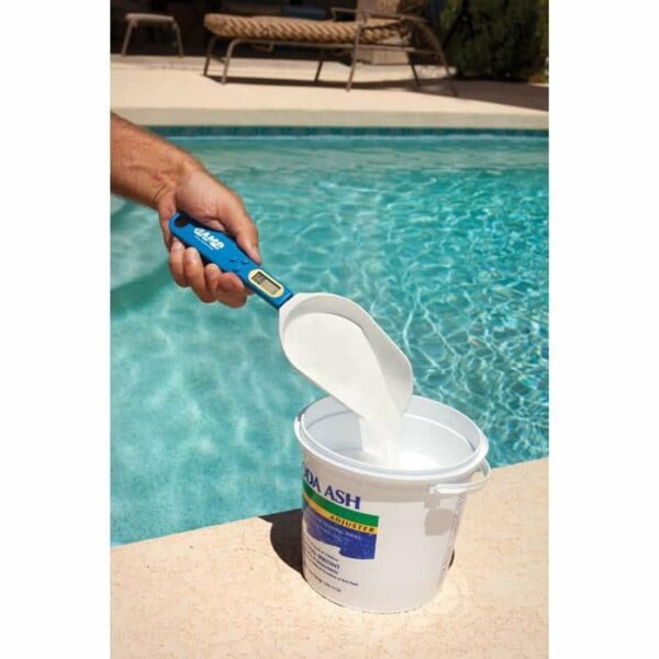 A hand uses the Accuweigh Chemical Scale & Scoop to measure white powder from a Soda Ash container by a swimming pool.