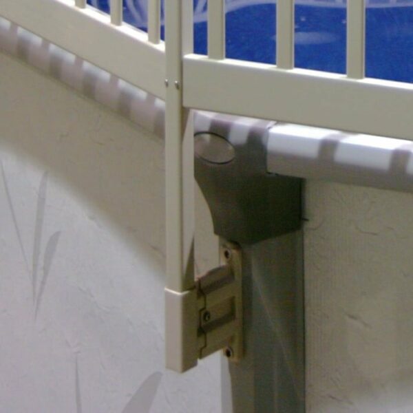 Close-up of the FENCE AG 24IN RESIN A WHITE VW baby safety gate, mounted on a wall with a metal bracket above patterned wallpaper near a blue background.