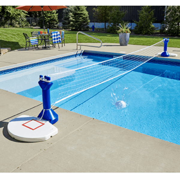 The Dunk & Spike 2in1 Combo is set up across a backyard pool, with a volleyball making a splash as it hits the water. Blue and white patio tables and chairs sit nearby, surrounded by lush trees and plants in the background.