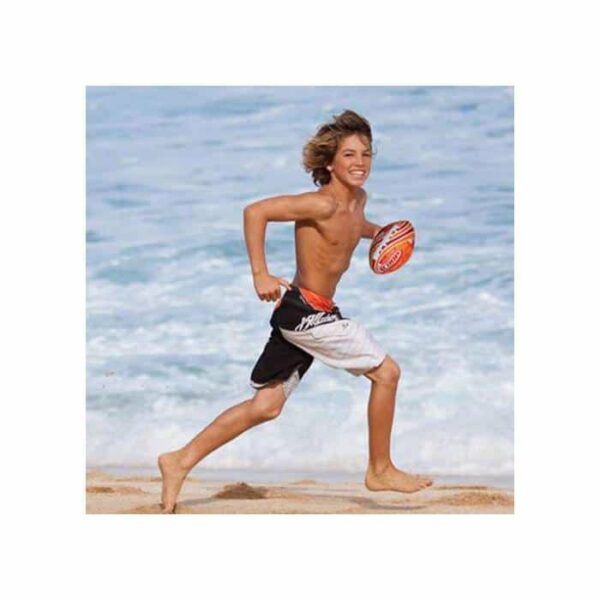 A smiling boy in swim trunks runs barefoot on the sandy beach while holding a Hydro Football, with ocean waves in the background.