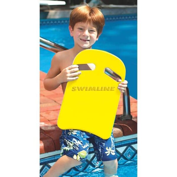 A smiling boy with reddish-brown hair in blue floral swim trunks stands by a pool, holding the Aqua-Coach Foam Kickboard—a bright yellow board with two hand grips.
