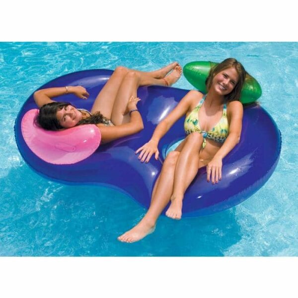 Two women in swimsuits relax on the SideBySide™ Lounger, a large blue and purple double pool float with headrests, as they float together in a clear swimming pool, smiling and enjoying the sunny day.