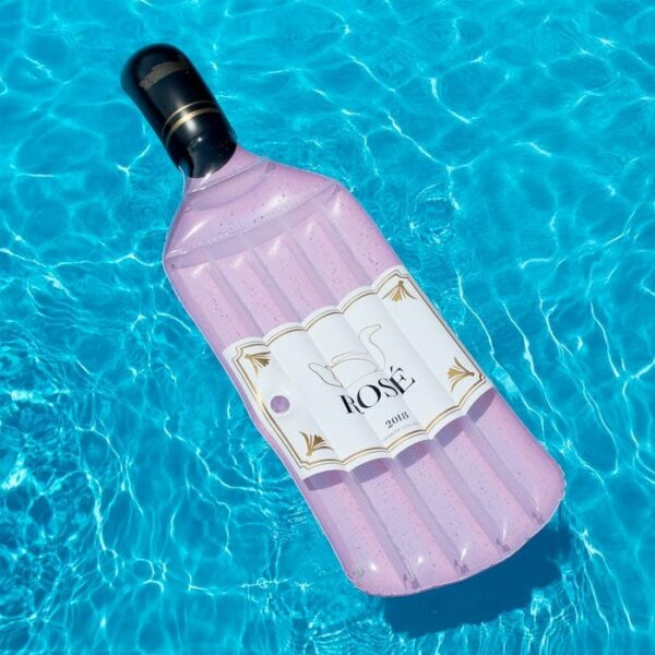 The Rose Float, a pink inflatable pool float shaped like a rosé wine bottle with a white and gold label, floats on the bright blue water of a swimming pool.
