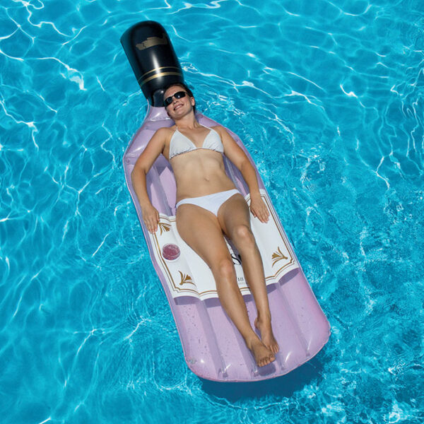 A woman in a white bikini and sunglasses lounges on The Rose Float, a large inflatable shaped like a purple champagne bottle, drifting in a clear blue swimming pool.