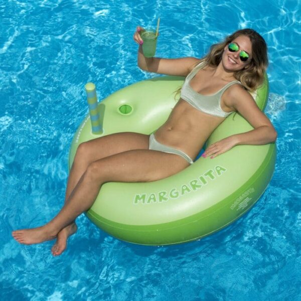 A woman relaxes and smiles on the MARGARITA RING, a green inflatable pool float shaped like a margarita, while holding a drink in a bright blue swimming pool.
