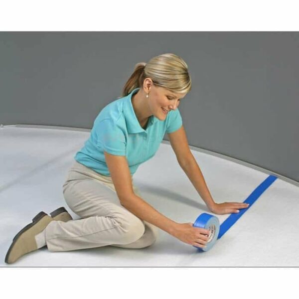 A woman in a light blue polo and beige pants kneels, applying Sure Seam™ Tape Roll (2 x 180) along a white surface near a gray wall.