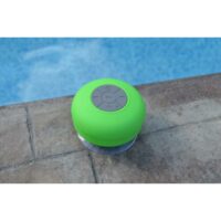 The SOUNDWAVES-OUTDOOR SPEAKER, a green waterproof Bluetooth device with gray control buttons, rests on a stone pool deck by clear blue water.