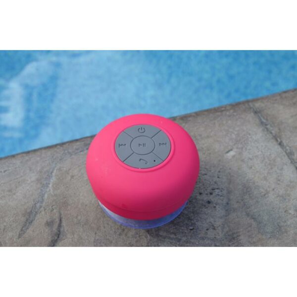 A SOUNDWAVES-OUTDOOR SPEAKER, in pink and waterproof, rests on a stone ledge by the blue water of a swimming pool.