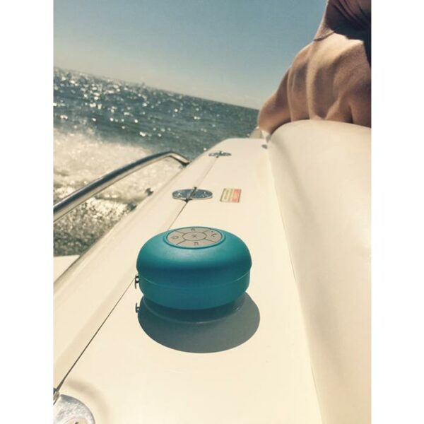 A SOUNDWAVES-OUTDOOR SPEAKER sits on a white boat surface under sunlight, with waves in the background and part of a persons arm visible.