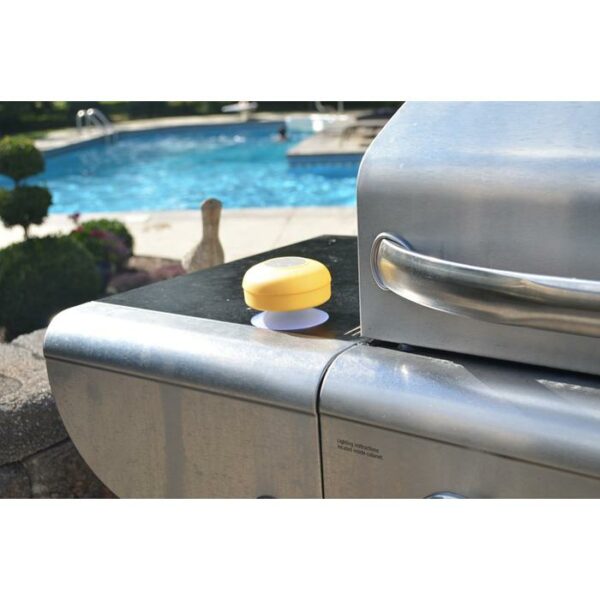 A yellow SOUNDWAVES-OUTDOOR SPEAKER rests on the side shelf of a stainless steel outdoor grill near a swimming pool, with shrubs and poolside decor in the background.