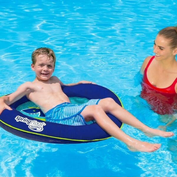 A smiling boy relaxes on the Spring Float Kids Boat in a clear, sunlit pool while a woman in a red swimsuit stands nearby, watching over him.