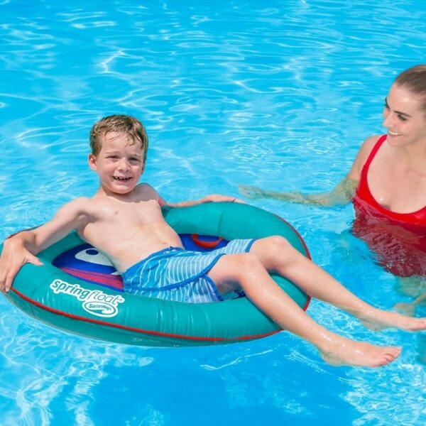 A smiling boy relaxes on the Spring Float Kids Boat in a clear blue swimming pool while a woman in a red swimsuit stands nearby.