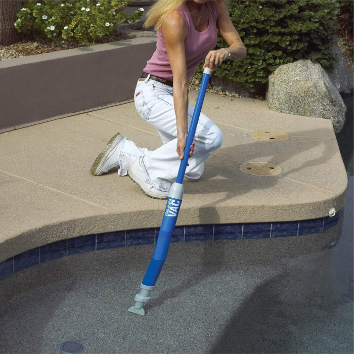 A person kneels beside a pool with tan paving and green shrubs, using the AQUA QUIK SPA/POOL VAC—a blue handheld vacuum—to clean the pool bottom.
