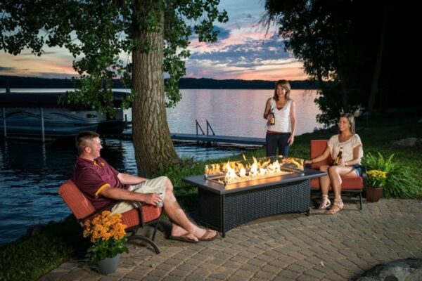 Three people relax by a lake at sunset around the Montego Firepit Table on a patio. Two women hold drinks while standing and sitting, as a man lounges nearby. Trees and a docked boat are visible in the background.