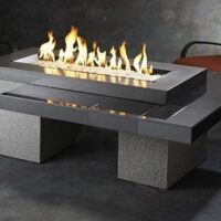 The Uptown Gas Firepit Table features a rectangular design with central flames, dark tile and stone accents, and is paired with two cushioned patio chairs in a softly lit setting.