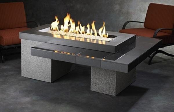 The Uptown Gas Firepit Table features a rectangular design with central flames, dark tile and stone accents, and is paired with two cushioned patio chairs in a softly lit setting.