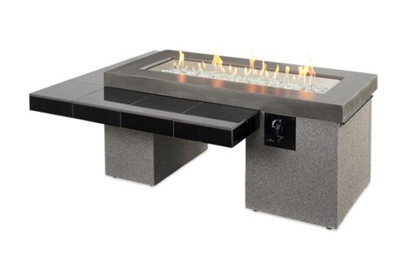 The Uptown Gas Firepit Table is a modern rectangular outdoor fire pit with a glass-enclosed gas flame in the center, featuring a grey stone base and black tiled side surface.