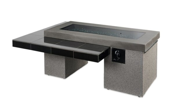 The Uptown Gas Firepit Table features a modern rectangular design with a concrete base, black tile side extension, control knob, and a glass-encased fire area filled with reflective glass stones.