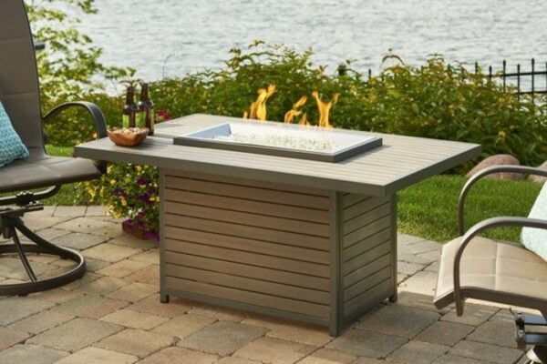 The Brooks Firepit Table features vibrant flames and is surrounded by patio chairs on a stone patio with green foliage and water in the background. Bottles and glasses are set on the tables edge for a relaxing outdoor experience.