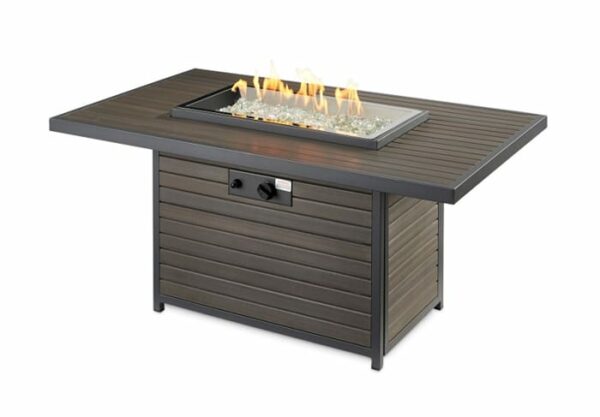 The Brooks Firepit Table features a rectangular outdoor gas design with a wood-like slatted surface, black trim, and a glass-covered fire area with decorative fire glass and visible flames. A front control knob and panel ensure easy operation.