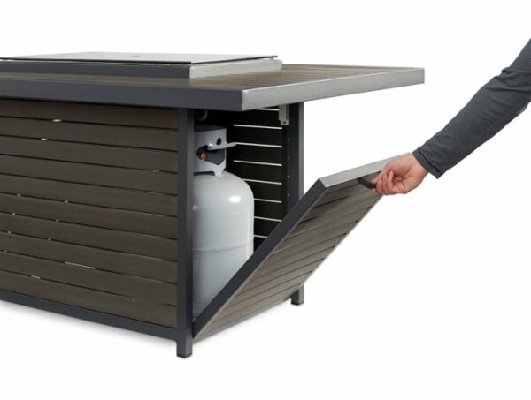 A person opens a slanted door on the Brooks Firepit Table, revealing a propane tank stored inside its base. The table features horizontal slats and a sturdy metal frame.