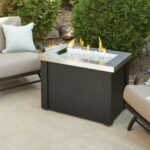 Two cushioned patio chairs with patterned pillows are arranged around the Providence Firepit Table, its flames glowing on a concrete patio bordered by greenery and rocks.