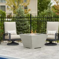 Two cushioned patio chairs with striped pillows face a Cove Sq Gas Firepit Bowl on a stone patio by the pool, surrounded by potted plants and lanterns, with a black metal fence and trees in the background.