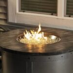 The Edison Firepit Table, featuring a dark wooden top and central flames, is placed on a patio near a window and chair.