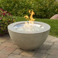 The Cove Gas Firepit Bowl 20 with glass stones is lit on a paved patio, surrounded by grass, flowering plants, and large rocks in the background.