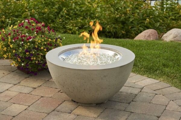 The Cove Gas Firepit Bowl 20 with glass stones is lit on a paved patio, surrounded by grass, flowering plants, and large rocks in the background.
