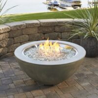 The Cove Gas Fire Pit Bowl 30 with glass rocks glows on a stone patio, framed by a curved stone wall and potted plants, while a lake with boats can be seen in the background.