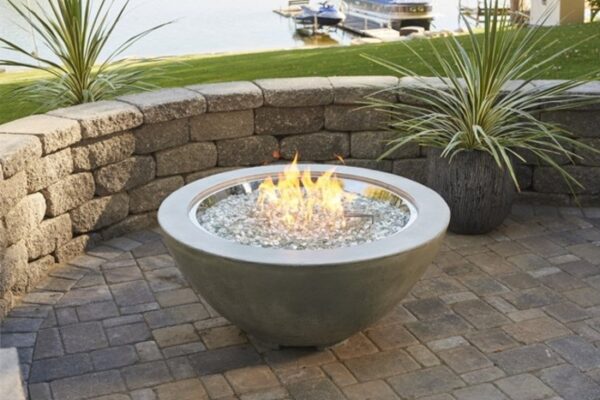 The Cove Gas Fire Pit Bowl 30 with glass rocks glows on a stone patio, framed by a curved stone wall and potted plants, while a lake with boats can be seen in the background.