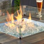 A 24x24 square tempered glass wind guard surrounds a fire pit table with burning flames over clear stones, beer glasses and bottles on the tabletop.