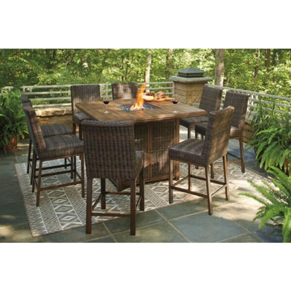 The Paradise Trail Bar Set (9 pc) features a square wooden fire pit table and six wicker bar-height chairs on a patterned rug, all set on a stone tile patio with lush green trees and plants in the background.
