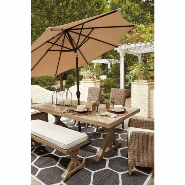 The Beachcroft 6-Piece Dining Group features a wooden table, cushioned bench, wicker chairs, tan umbrella, and hexagonal-patterned rug. Set outdoors near white brick walls and greenery, the table is decorated with plates and decor.