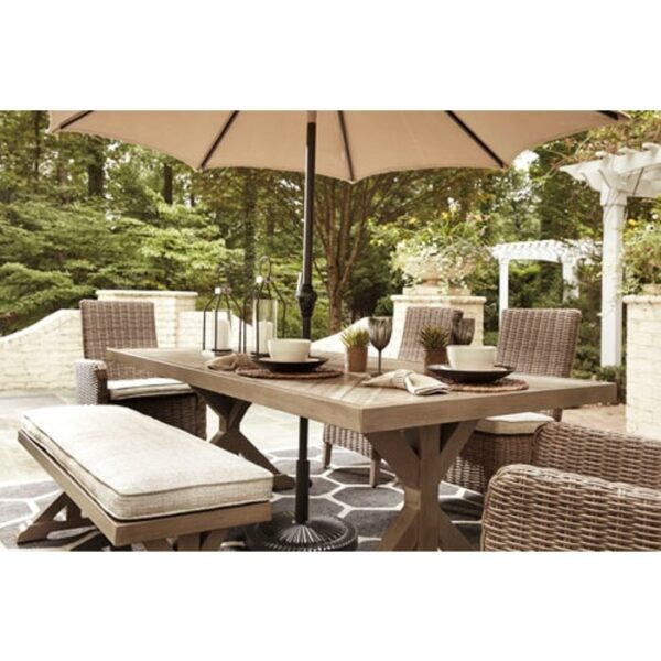 The Beachcroft 6-Piece Dining Group features a wooden table, wicker chairs, a cushioned bench, and a large beige umbrella. Surrounded by greenery and white brick walls, the table is elegantly set with plates and decor for outdoor dining.