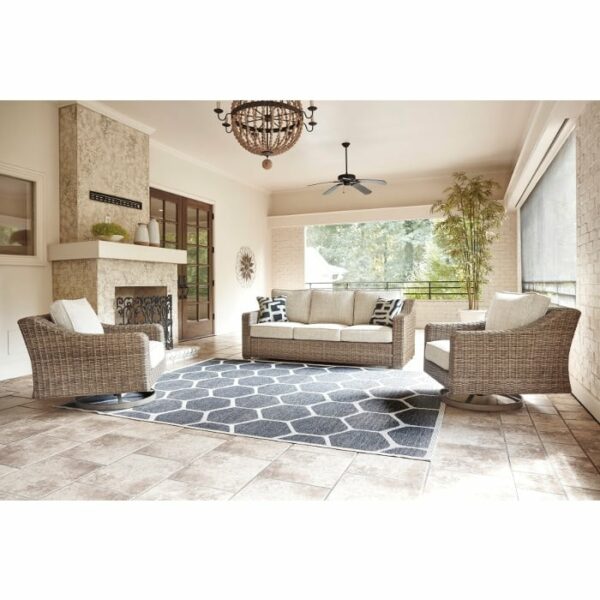 A bright sunroom features the Beachcroft Deep Seating Group—wicker sofa and armchairs with neutral cushions—a geometric rug, fireplace, potted plants, and large windows flooding the room with natural light.