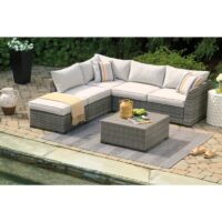 The Cherry Point Sectional (4 pc) features a gray wicker sofa with beige cushions, striped pillows, and a matching coffee table on a gray rug by the pool. A yellow blanket, drink tray, and plants complete the inviting patio setup.