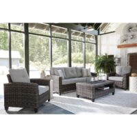 A modern sunroom with large windows showcases the Cloverbrooke Grouping 4 piece set—wicker furniture with light gray cushions, a matching coffee table, a patterned rug, and potted plants against green trees outside.