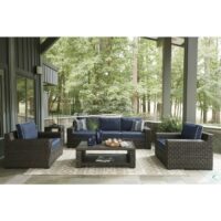 The Grasson Lane 5-Piece Grouping features a wicker sofa, two armchairs, and a glass-top coffee table on a patterned rug with blue cushions and white piping. Set on a covered porch with gray columns, it’s surrounded by lush trees and greenery.