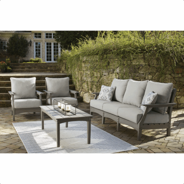 Create an inviting outdoor space with the Visola Lounge Grouping 4 piece, featuring a gray sofa, two matching armchairs, and a rectangular coffee table on a stone patio with lush greenery and a stone wall backdrop.