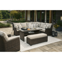 The Brook Ranch Grouping 4-Piece features a wicker sectional sofa, bench, and armchair with beige cushions, a rectangular coffee table, striped and patterned pillows, plus a white rug on a stone patio with greenery and a building in the background.