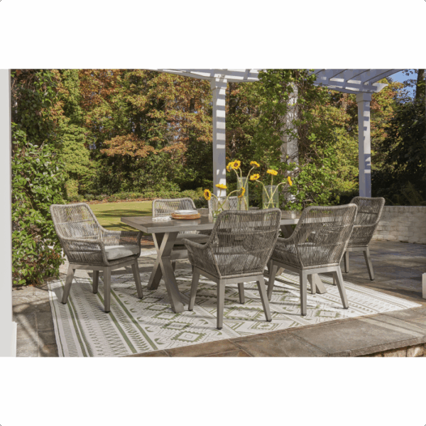 The Beach Front Wicker Set 7 piece features a rectangular table, six woven chairs, and a stylish area rug, arranged outdoors under a pergola with greenery and trees in the background.
