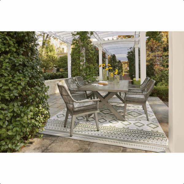 The Beach Front Wicker Set 7 piece features a rectangular table, six woven chairs, and a vase of sunflowers atop a patterned rug under a pergola, surrounded by lush greenery and a stone patio for stylish outdoor dining.