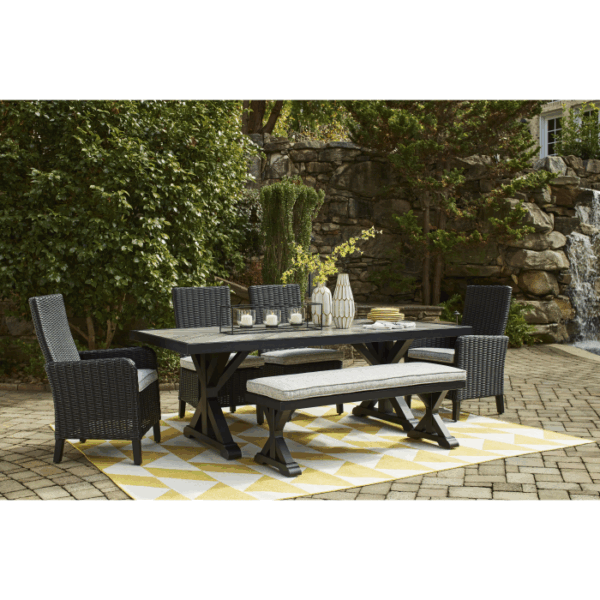 The Beachcroft Dining Set Black 6 piece features a dark table, four woven chairs, a bench, and a yellow and white geometric rug on a stone patio. The table is styled with candles and vases, surrounded by greenery and a rock waterfall background.