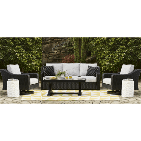 The Beachcroft Deep Seating Group Black 4 piece features a gray cushioned sofa, two armchairs, and a black coffee table on a yellow and white rug, accented by white side tables amid greenery and stone walls.
