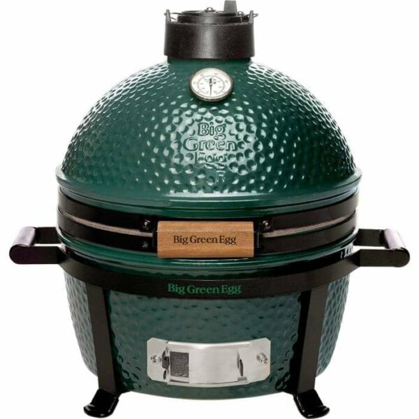 The Big Green Egg MiniMax is a green, dome-shaped ceramic grill with a temperature gauge on top, sturdy black metal legs and side handles, and a wooden handle.