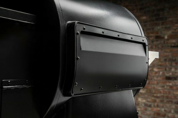 Close-up of a Timberline 1300 Pellet Grill in black metal, positioned against a brick wall, featuring the side firebox with visible bolts and a rectangular access panel.