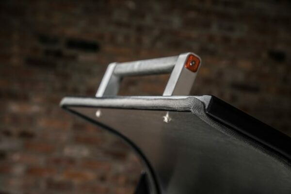 Close-up of the Timberline 1300 Pellet Grill, showcasing its grey fabric edge, metallic handle, and visible screws against a blurred brick wall background.