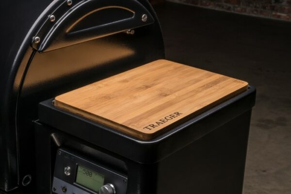 A close-up of the Timberline 1300 Pellet Grill with a rectangular wooden cutting board on top, featuring TRAEGER engraved in the corner. The grill is indoors against a dark background.